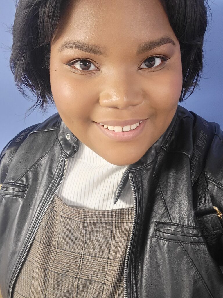 A brown skinned woman with chin length hair stands in front of an indigo colored wall. She is framed in photo from the chest up, smiling for a selfie.