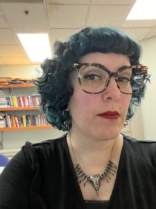 Femme-presenting person with navy curly hair and large-framed glasses sits in front of a pale wall with a bookshelf.