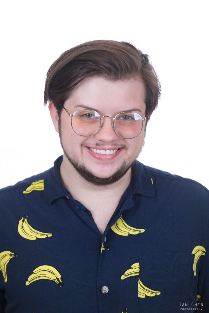 This photo is a headshot of the editor, who is a nonbinary person with short, dark brown hair, a closely-trimmed beard, and a very faint mustache. They are looking directly at the camera and smiling, wearing circle glasses that cast a peach color over their eyes. They are wearing a navy blue button-up shirt with a bright yellow banana pattern.